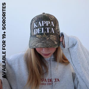 May include: A gray hoodie with the text "Kappa Delta since 1897" on the front. The person is wearing a camouflage baseball cap with the text "Kappa Delta" in white letters.