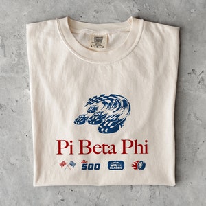 Puede incluir: Una camiseta blanca con un gráfico azul y negro de un coche de carreras. El texto "Pi Beta Phi" está impreso en rojo debajo del gráfico. El texto "the 500" y "CLASSIC" están impresos en negro debajo del texto "Pi Beta Phi".