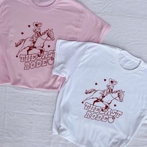 May include: Two t-shirts with a cowgirl riding a horse and the text "The Last Rodeo" printed on them. The shirt on the left is pink and the shirt on the right is white. The white shirt has a diamond ring symbol next to the text.