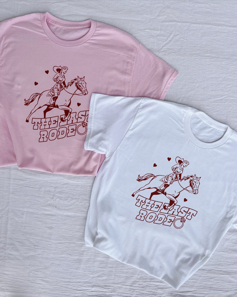 May include: Two t-shirts with a cowgirl riding a horse and the text "The Last Rodeo" printed on them. The shirt on the left is pink and the shirt on the right is white. The white shirt has a diamond ring symbol next to the text.