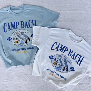 Op de afbeelding: Twee T-shirts met een grafisch ontwerp van een berglandschap met een tent, een rivier en bomen. De tekst "CAMP BACH" is in blauw op beide shirts gedrukt. De tekst "THE LAST TRAIL BEFORE THE VEIL" is onder de afbeelding gedrukt. Het shirt links is lichtblauw en het shirt rechts is wit.