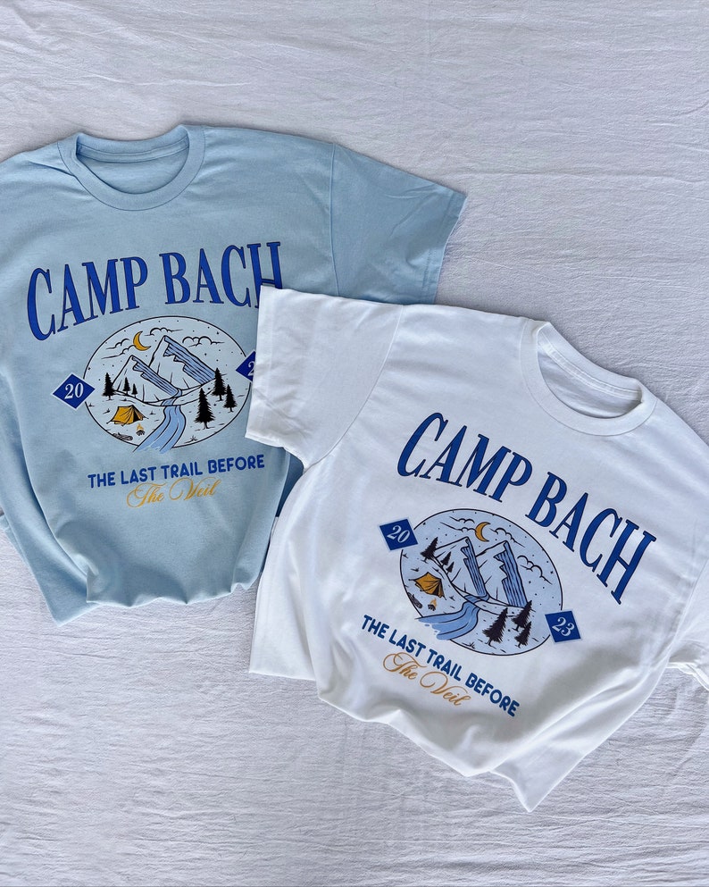 Op de afbeelding: Twee T-shirts met een grafisch ontwerp van een berglandschap met een tent, een rivier en bomen. De tekst "CAMP BACH" is in blauw op beide shirts gedrukt. De tekst "THE LAST TRAIL BEFORE THE VEIL" is onder de afbeelding gedrukt. Het shirt links is lichtblauw en het shirt rechts is wit.