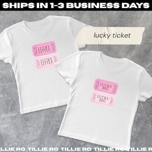 May include: Two white t-shirts with pink ticket graphics that read "LUCKY LITTLE" and "LUCKY BIG". The text "lucky ticket" is above the shirts. The shirts are short-sleeved.