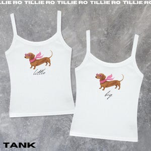 May include: Two white tank tops with spaghetti straps. Each top features a brown dachshund wearing pink sunglasses and a scarf. One says "little" and the other says "big" in script.