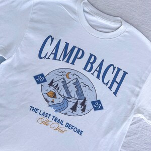Op de afbeelding: Wit T-shirt met een blauw grafisch ontwerp met de tekst "Camp Bach" en een berglandschap met een tent, een rivier en bomen. De grafiek bevat ook de tekst "The Last Trail Before The Veil" en de nummers "20" en "23".