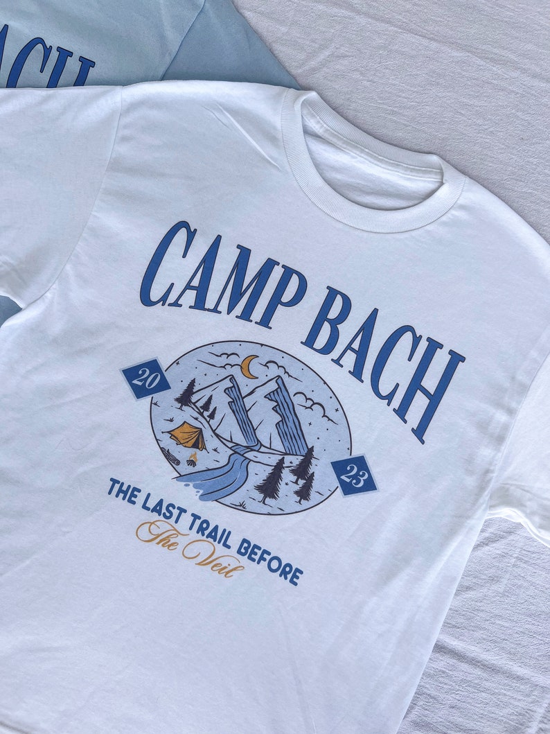 Op de afbeelding: Wit T-shirt met een blauw grafisch ontwerp met de tekst "Camp Bach" en een berglandschap met een tent, een rivier en bomen. De grafiek bevat ook de tekst "The Last Trail Before The Veil" en de nummers "20" en "23".