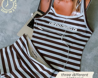 May include: A brown and light blue striped tank top with the text alpha delta pi printed on the chest, paired with matching striped shorts. The tank top features thin straps and a square neckline, with white wired earbuds resting on the fabric.