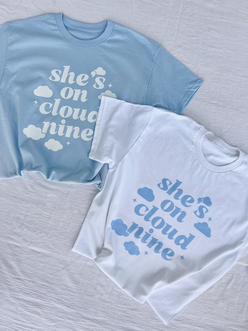May include: Two t-shirts with the text "she's on cloud nine" printed on them. The shirts are light blue and white with blue clouds and stars.