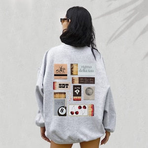 Puede incluir: Sudadera gris con un collage de etiquetas de cajas de cerillas en la espalda. Las etiquetas muestran imágenes de un vaquero montando un caballo, una bola de billar, cerezas y el texto "sigma delta tau" y "SDT".