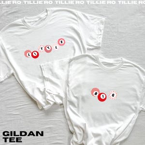 May include: Two white Gildan brand t-shirts. One shirt has the word "LITTLE" in red and pink circles. The other shirt has the word "BIG" in red and pink circles. The shirts are on a white background.