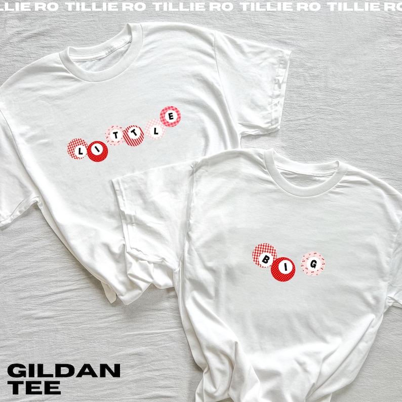 May include: Two white Gildan brand t-shirts. One shirt has the word "LITTLE" in red and pink circles. The other shirt has the word "BIG" in red and pink circles. The shirts are on a white background.