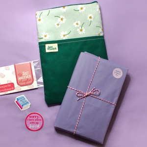 May include: A green and white daisy-patterned book sleeve, a wrapped book tied with red and white twine, a magnetic bookmark, and stickers with book-related phrases on a purple background.