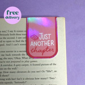 Pode incluir: Um marcador de página rosa e vermelho com acabamento holográfico. O marcador de página tem o texto "Just another chapter" com um pequeno ícone de livro.