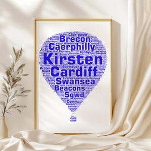 Könnte beinhalten: Ein gerahmter Druck mit einem blauen Heißluftballon-Design, das mit Ortsnamen in Weiß gefüllt ist. Das Wort "Kirsten" ist prominent dargestellt. Weitere sichtbare Wörter sind "Cardiff", "Swansea" und "Brecon". Der Druck wird vor einem cremefarbenen Hintergrund präsentiert.