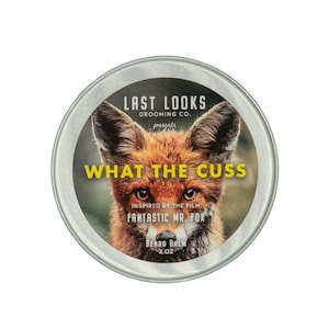 May include: A silver tin with a yellow label that reads "Last Looks Grooming Co. presents What The Cuss" and features a fox with the text "Inspired by the film Fantastic Mr. Fox Beard Balm 2 oz."
