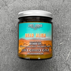 May include: A glass jar with a silver lid, labelled "DEAR ALIEN" with a desert landscape graphic. The label also includes the text "LAST LOOKS GROOMING CO." and "ASTEROID CITY". The jar contains approximately 227 grams of soy wax.