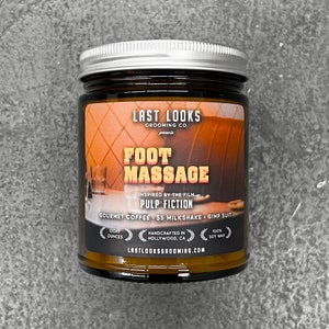 May include: A brown glass jar with a silver lid. The label reads "LAST LOOKS GROOMING CO. presents FOOT MASSAGE INSPIRED BY THE FILM PULP FICTION GOURMET COFFEE $5 MILKSHAKE GIMP SUIT EIGHT OUNCES HANDCRAFTED IN HOLLYWOOD, CA 100% SOY WAX LASTLOOKSGROOMING.COM".