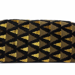 May include: A rectangular toiletry bag with a geometric pattern of black and gold triangles on a brown background. The bag has a black leather handle on one side and a black leather loop on the other.