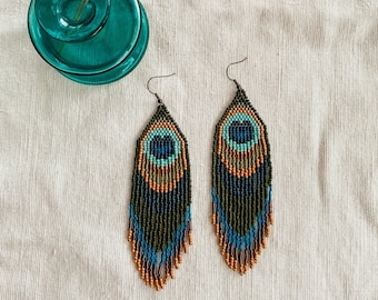 May include: A pair of long, beaded earrings with a peacock feather design. The earrings are made with green, blue, and gold beads.