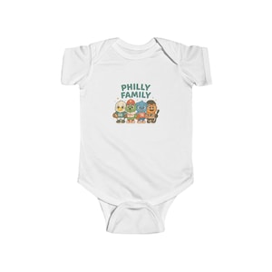 May include: A white short-sleeve baby bodysuit featuring a graphic print of four cartoon mascots in sports uniforms. Above the characters, the text reads PHILLY FAMILY in green letters. The mascots are depicted in various colors including white, green, blue, and orange.
