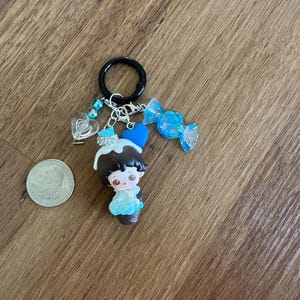 May include: A handmade keychain featuring a small figurine with dark hair and a blue dress, adorned with a blue heart, a candy charm, and a clear heart charm. The keychain is attached to a black ring.
