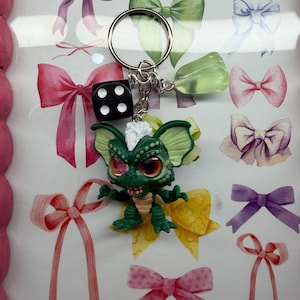 May include: A keychain featuring a green creature figurine with large orange eyes and a white tuft of hair, accompanied by a black six-sided die with white pips and a translucent light green geometric charm attached to a silver metal ring.