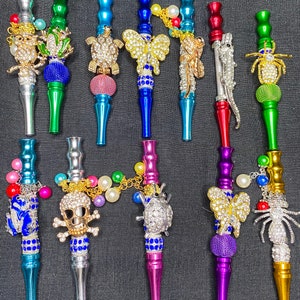 May include: Ten colorful metal smoking pipes with decorative charms. The charms include a butterfly, a turtle, a skull, a spider, a frog, a ladybug, an alligator, and a dragonfly. The pipes are decorated with rhinestones and beads.