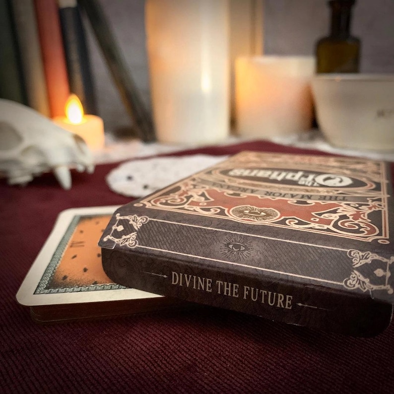 May include: A close-up of a tarot card deck box and a few cards. The box is brown with ornate gold and red designs and the text "Orphans The Major Arcana" and "Divine the Future". The cards have an orange and white design.