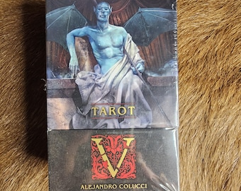 May include: A tarot card deck featuring a blue-skinned figure with bat-like wings, holding a sword. The word "TAROT" is printed in gold. The bottom of the box has a red and gold design with the name "ALEJANDRO COLUCCI".