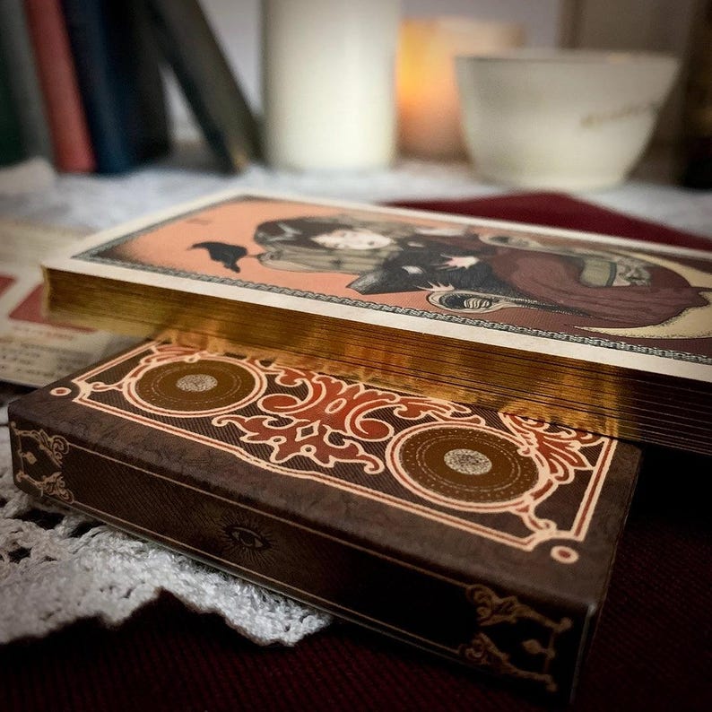 May include: A deck of tarot cards with gold gilded edges and a decorative box. The box is brown with an ornate design in red and gold. The top card features an illustration of a person in a cloak.
