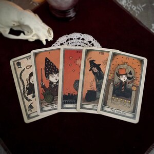 May include: Five tarot cards with illustrations of figures in a mystical theme. The cards feature images of a wizard, a witch, and other characters. The cards are arranged on a dark red surface with a skull and lace doily in the background.