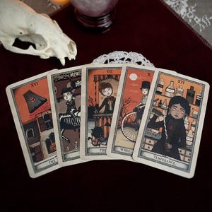 May include: Five tarot cards with gothic illustrations. Each card features a unique design with characters, objects, and numbers. The cards have an orange and cream color scheme, with the words "The", "Wheel", and "Temperance" visible.