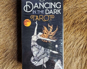 May include: A black tarot card box with the words "Dancing in the Dark Tarot" in white and gold lettering. The box features an illustration of a woman dancing with a full moon and a wolf.