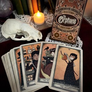 May include: A tarot card deck with the title "The Orphans" and the words "Divine the Future" on the box. Several cards are fanned out, each with unique illustrations and titles like "The Hermit" and "The High Priestess". A skull and candle are also present.