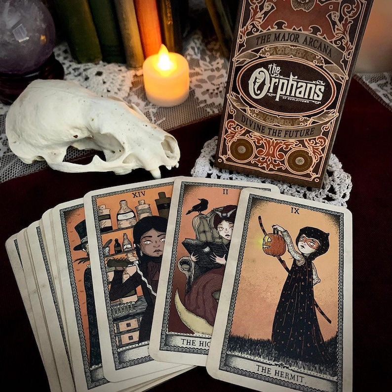 May include: A tarot card deck with the title "The Orphans" and the words "Divine the Future" on the box. Several cards are fanned out, each with unique illustrations and titles like "The Hermit" and "The High Priestess". A skull and candle are also present.