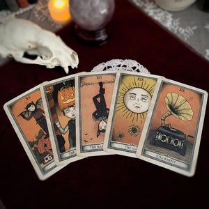 May include: Five tarot cards with vintage illustrations are displayed. The cards feature images of a bat-like figure, a person with a pumpkin head, a figure hanging upside down, a sun with a face, and a vintage gramophone. The cards are set against a dark red background.