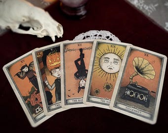May include: Five tarot cards with vintage illustrations are displayed. The cards feature images of a bat-like figure, a person with a pumpkin head, a figure hanging upside down, a sun with a face, and a vintage gramophone. The cards are set against a dark red background.