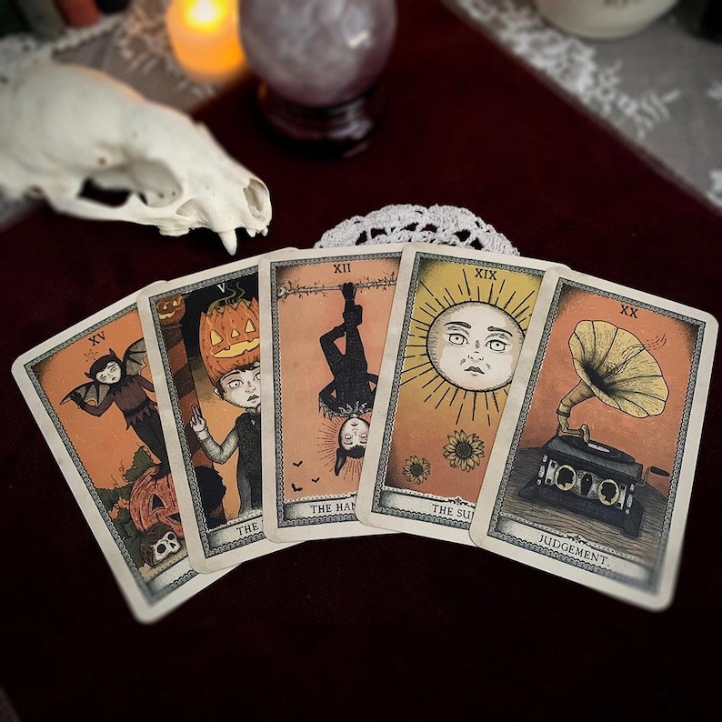 May include: Five tarot cards with vintage illustrations are displayed. The cards feature images of a bat-like figure, a person with a pumpkin head, a figure hanging upside down, a sun with a face, and a vintage gramophone. The cards are set against a dark red background.