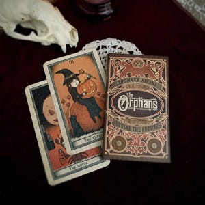 May include: A tarot card deck with a brown box featuring the text "The Major Arcana" and "The Orphan's". Two tarot cards are visible, one with a witch holding a pumpkin. The cards have an orange and black color scheme, with a vintage aesthetic.