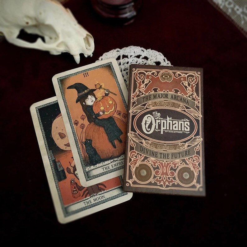 May include: A tarot card deck with a brown box featuring the text "The Major Arcana" and "The Orphan's". Two tarot cards are visible, one with a witch holding a pumpkin. The cards have an orange and black color scheme, with a vintage aesthetic.