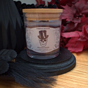 May include: A glass candle jar with a wooden lid. The candle is labeled "Undertaker" with a skull wearing a top hat and the text "Cerberus Treasure".