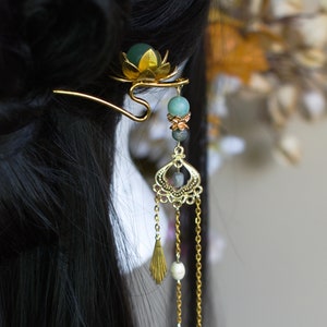 Op de afbeelding: Goudkleurige haarspeld met een lotusbloemdesign en bungelende kettingen. De speld heeft groene en witte kralen en waaiervormige bedels. Het haaraccessoire wordt in zwart haar gedragen.