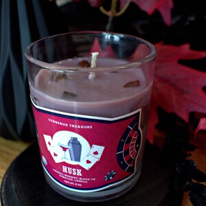 May include: A glass candle jar with a red, white, and blue label that says "Cerberus Treasure" and "Husk" with the scent description "Triball Whiskey, Black Tie Tobacco, Vanilla". The candle is a dark purple color and has a white wick.
