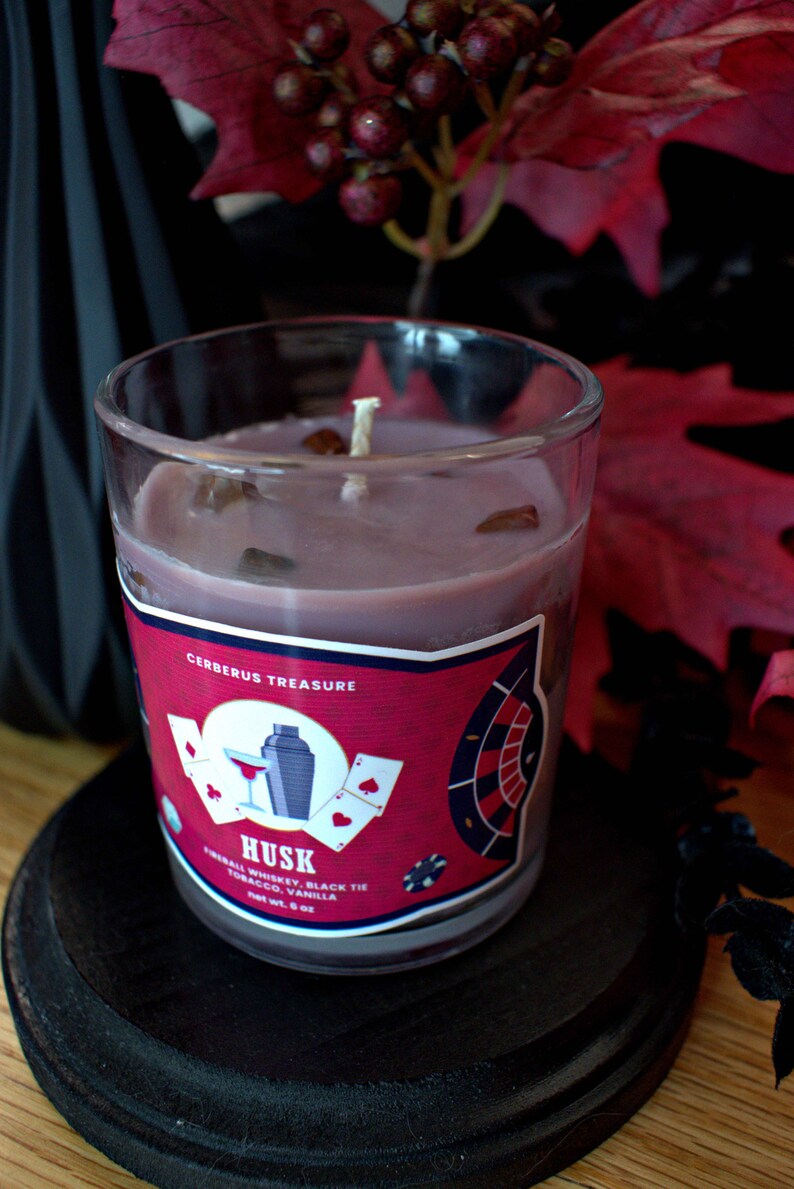 May include: A glass candle jar with a red, white, and blue label that says "Cerberus Treasure" and "Husk" with the scent description "Triball Whiskey, Black Tie Tobacco, Vanilla". The candle is a dark purple color and has a white wick.