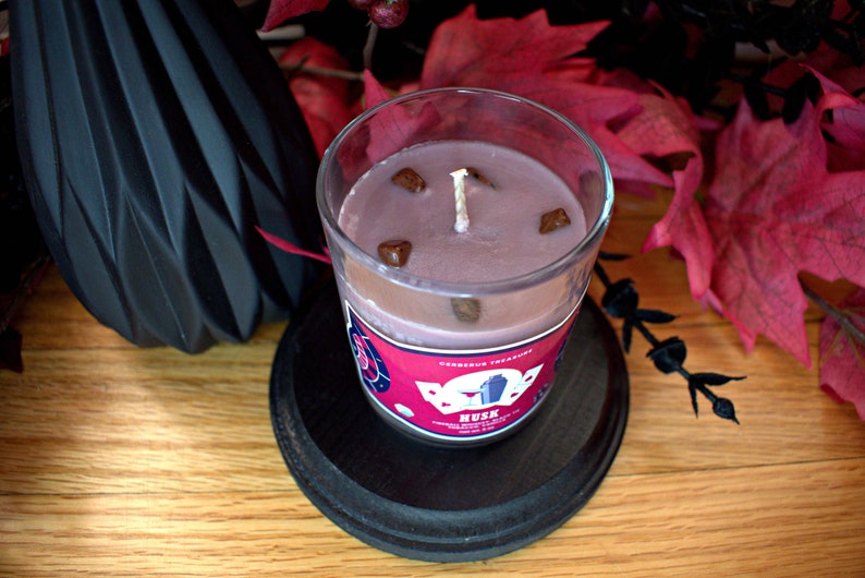 May include: A glass jar candle with a pink wax and brown decorative pieces. The label reads "Delicious Treasures" and "Musk" with a red and white design.