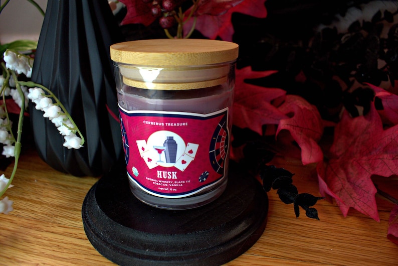 May include: A glass jar candle with a wooden lid. The label is red and black with the text "Cerberus Treasure" and "Husk" in white. The candle is labeled "Finest Whiskey, Black Tea, Tobacco, Vanilla" and is 8.5 oz.