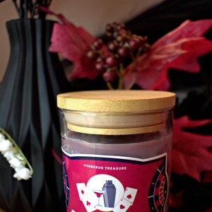 May include: A glass candle jar with a wooden lid. The label is red with black text and features a playing card design. The text on the label reads "Cerberus Treasure, Husk, Fireball Whiskey, Black Tie Tobacco, Vanilla, net wt. 6 oz."