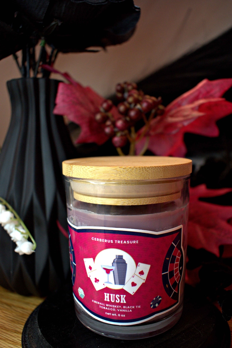 May include: A glass candle jar with a wooden lid. The label is red with black text and features a playing card design. The text on the label reads "Cerberus Treasure, Husk, Fireball Whiskey, Black Tie Tobacco, Vanilla, net wt. 6 oz."