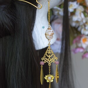 Op de afbeelding: Goudkleurig haaraccessoire met een lotusbloem en een hangend ontwerp. Het heeft een gestreepte steen, roze kralen en bladvormige hangers. Het haaraccessoire is versierd met een gouden ketting, wat een elegante uitstraling cre&euml;ert.