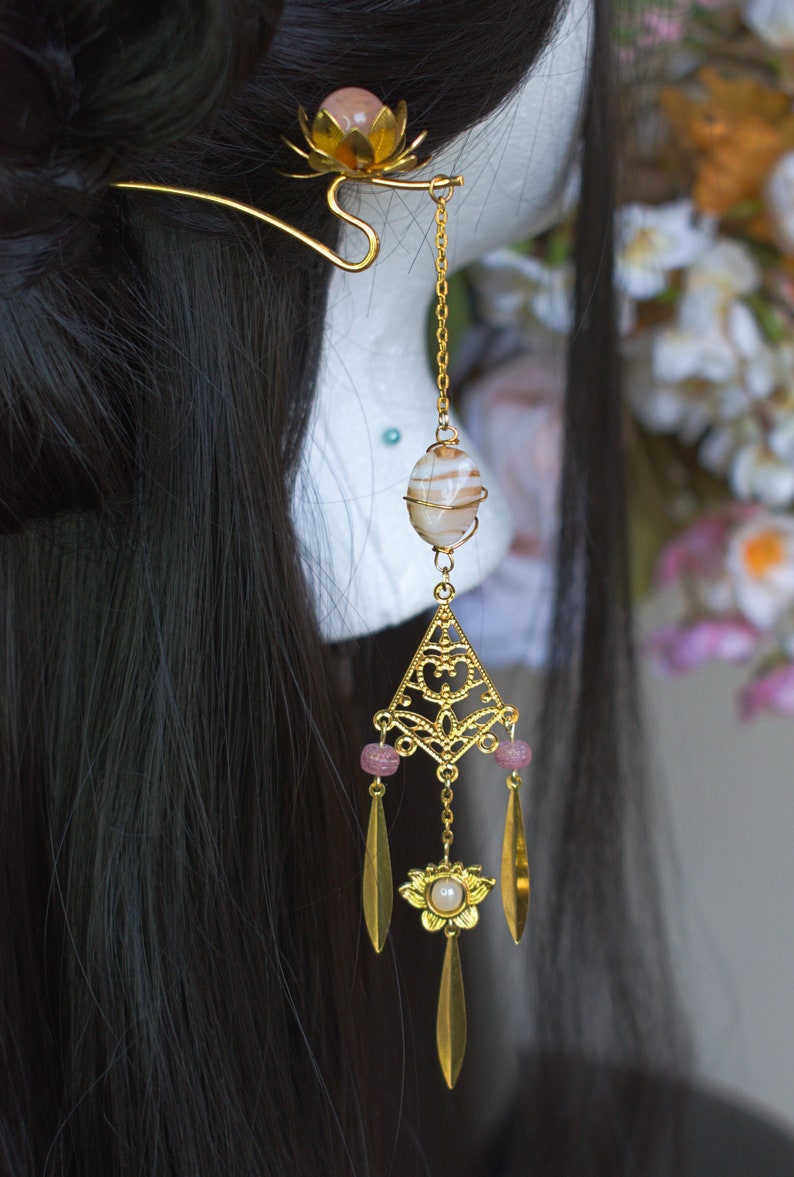 Op de afbeelding: Goudkleurig haaraccessoire met een lotusbloem en een hangend ontwerp. Het heeft een gestreepte steen, roze kralen en bladvormige hangers. Het haaraccessoire is versierd met een gouden ketting, wat een elegante uitstraling cre&euml;ert.